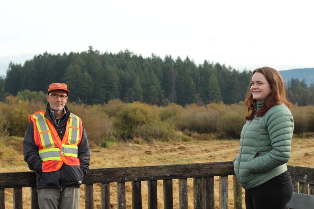 Paul Gowland stands to the left of the photo wearing an orange safety vest. Phaedra Douglass stands on the far right, facing sideways but looking at the camera, wearing a green puffy jacket. Behind them is wooden fencing from the boardwalk they're standing on and behind that is marshland, forest and mountain.