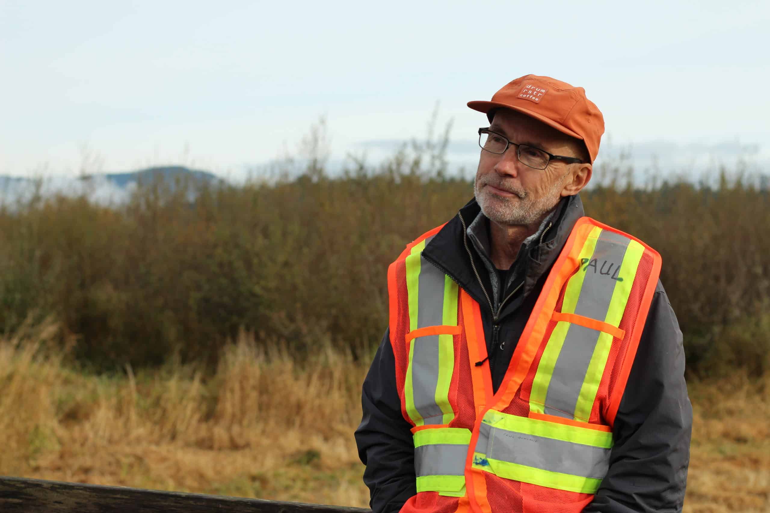 Paul Gowland stands on the left of the photo, facing the camera but looking beside it. He wears an orange safety vest and orange ball cap. Behind him is the marsh.