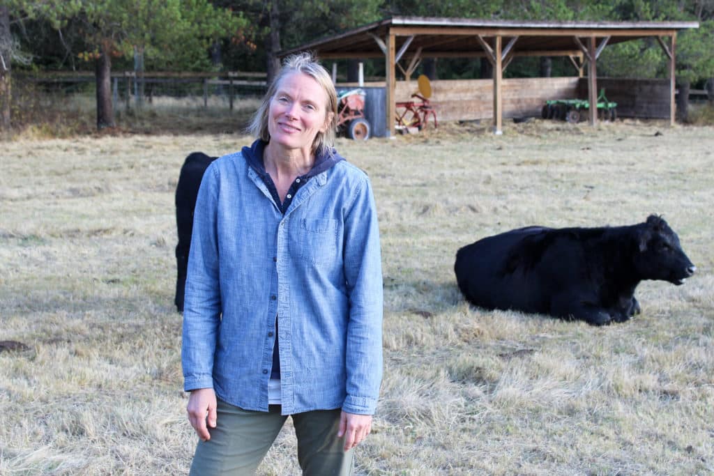 DeLisa Lewis stands in a field with a black cow laying down behind and to the right of her. A sliver of another black cow is seen behind her left side. She's wearing a sky blue jacket and smiling.