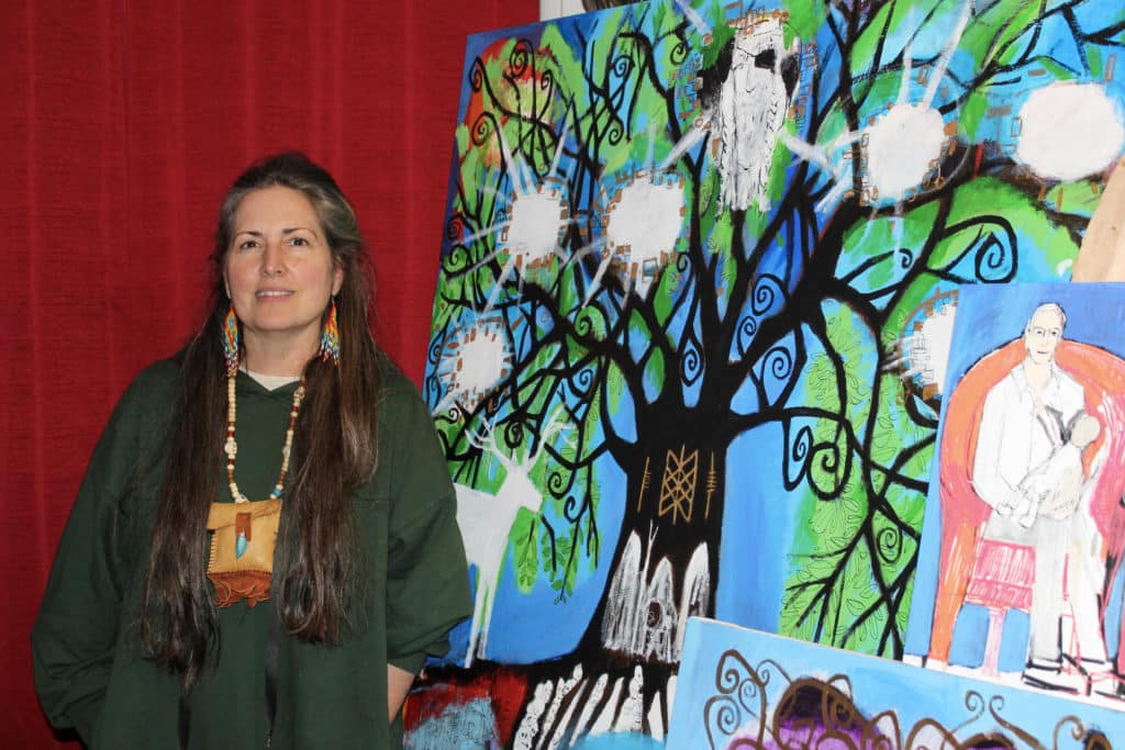 Artist Coco Jones stands in front of a red curtain and wears a green long-sleeve top. To the right of her is one of her paintings with a blue background. In the painting is a black tree with green leaves, white animals, norse symbols and a man sitting to the right of it holding a baby.