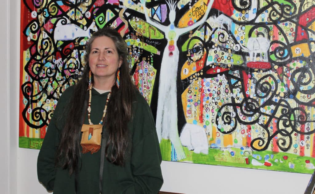 Coco Jones stands in front of a colourful painting with a tree and tall white figure standing in the middle of the tree with its arms stretch up. Jones wears a green long-sleeve top, long beaded earrings and a pouch around her neck.