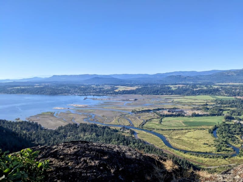 A view of Cowichan Bay as seen from atop a mountain. Blue water on the left meets green land and a river is seen flowing into the land. Hills are in the background.