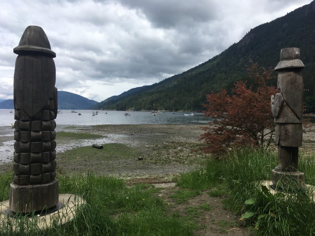 Two stone figures, their backs to the viewer and facing the ocean, are on either side of the photo. The figures are tall. In between, a view of the beach and ocean can be seen as well as the grass before the sandy shoreline. In the background of the photo is a large mountain that takes up the right side of it and a smaller-looking mountain further away on the left. There are clouds in the sky and the photo shows a cloudy day.