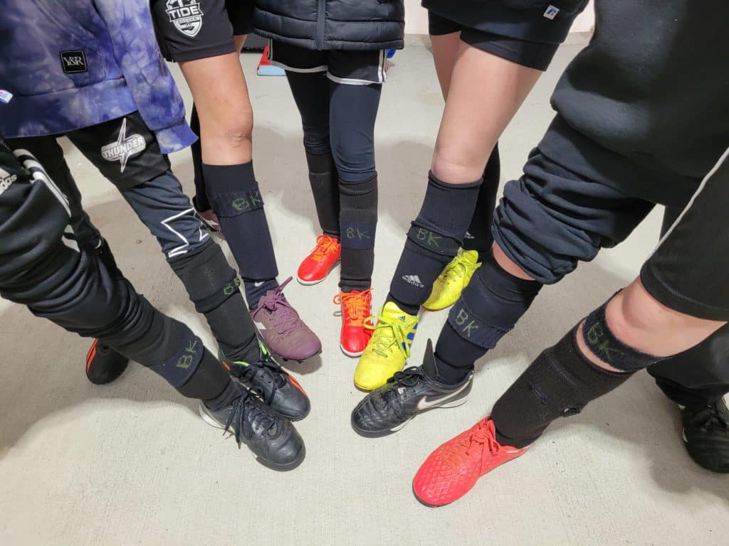 The photo is taken from above, showing the legs of seven soccer players pointing their feet into a circle. They're wearing their soccer uniform, with cleats, shin-pads and socks and black bands that don the initials BK for Bill Keserich Junior.