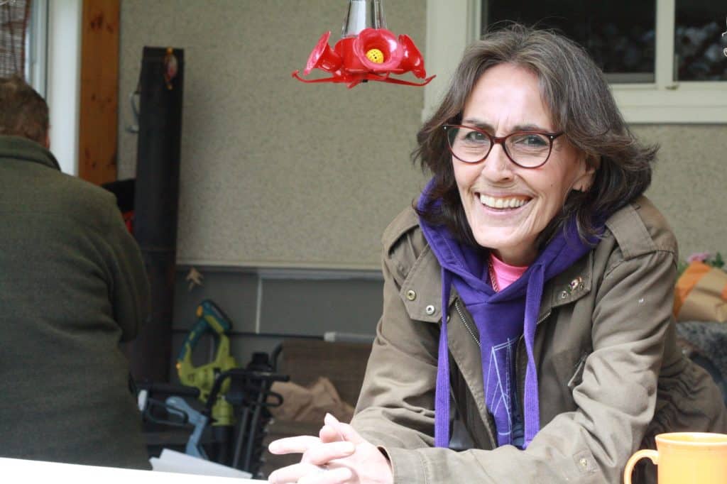 A woman smiles with hands on clasped on a table, hummingbird feeder in background.