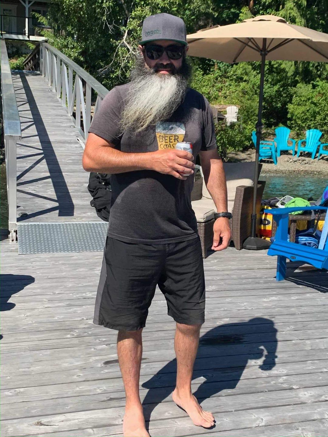 Bill Keserich Jr. stands on a dock, barefoot, holding a beer. His beard is long and grey and he wears black shorts and a faded black t-shirt.