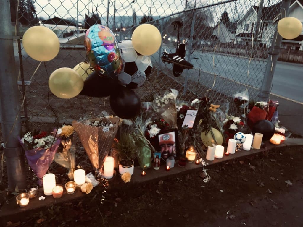 A line of lit candles are on the ground in front of a fence that surrounds the soccer field. Flowers are placed behind and around the candles and balloons and cleats are hung on the fence above the candles and flowers. A photo of Bill Keserich Jr. is seen as well.