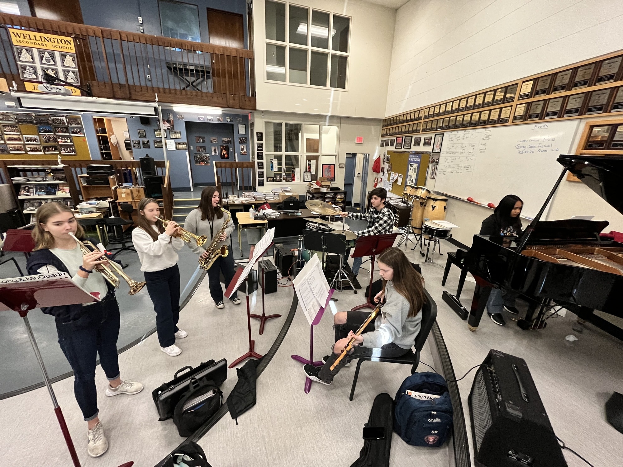 Six students play various instruments in a large music classroom. Three studnets standing on the left side of the photo, facing to the right of the photographer, play brass instruments. In the center of the picture, facing left, a student plays drums and another plays electric bass. To the right of the photo a student sits at a grand piano facing the piano music stand.