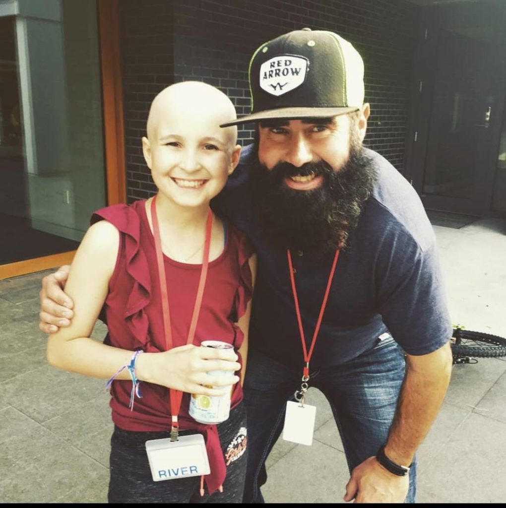 Poppy White stands on the left, wearing a red shirt. Her head is bald and she has a smile on her face. Bill Keserich Junior is on her right, hunched over with his hand around her shoulders and smiling. He wears a grey shirt, jeans and black ball cap. His beard is lengthy and black.