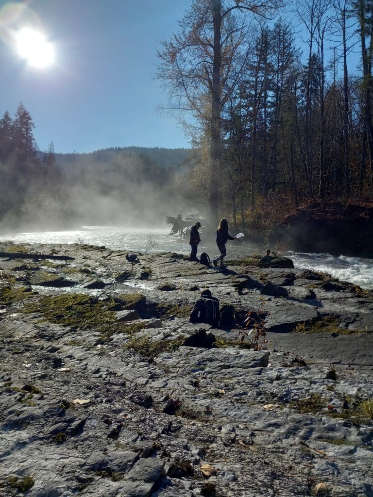 A wide river bank leading to the river which has mist coming off it. The sun shines in the top left corner of the photo surrounded by blue sky and rays of sun peer through the mist. In the distance the river bends and is surrounded by trees with mountains seen in the far distance. Silhouettes of students walking and sitting along the river are on the river bank closest to the photographer in this image.