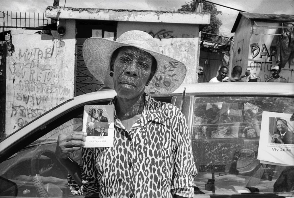 A black and white photo of a Haitian woman wearing a weaved sunhat that casts a checkered shadow over her face. She looks pensively at the camera, head tilted to the side. In her right hand is a campaign pamphlet for the 2001 Haiti elections