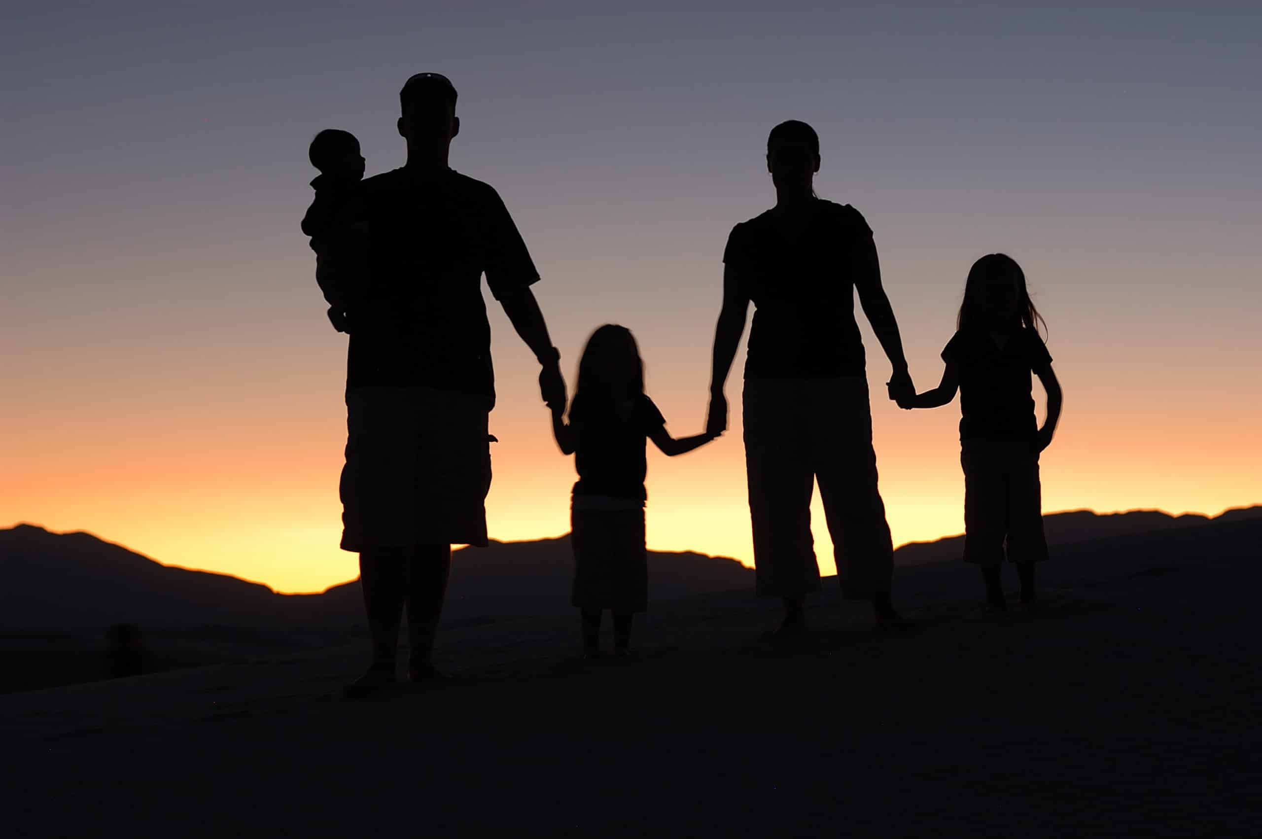 The silhouette of a parent, child, parent and another child standing in a line and holding hands with their backs to the camera, facing a sunset.
