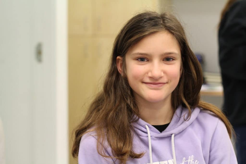 A photo of Mill Bay Nature School student Vienna Wright, taken from the shoulders up. She is smiling and has long brunette hair. She is wearing a lavender coloured hoodie. The blurred background behind her is of her classroom.