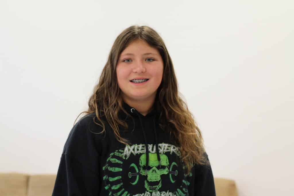 A photo of Juna Murray, taken from the chest-up. She is smiling and has long, curly, brunette hair. She is wearing a black hoodie with a neon green skull in the centre and the words "axel busters" above the skull.