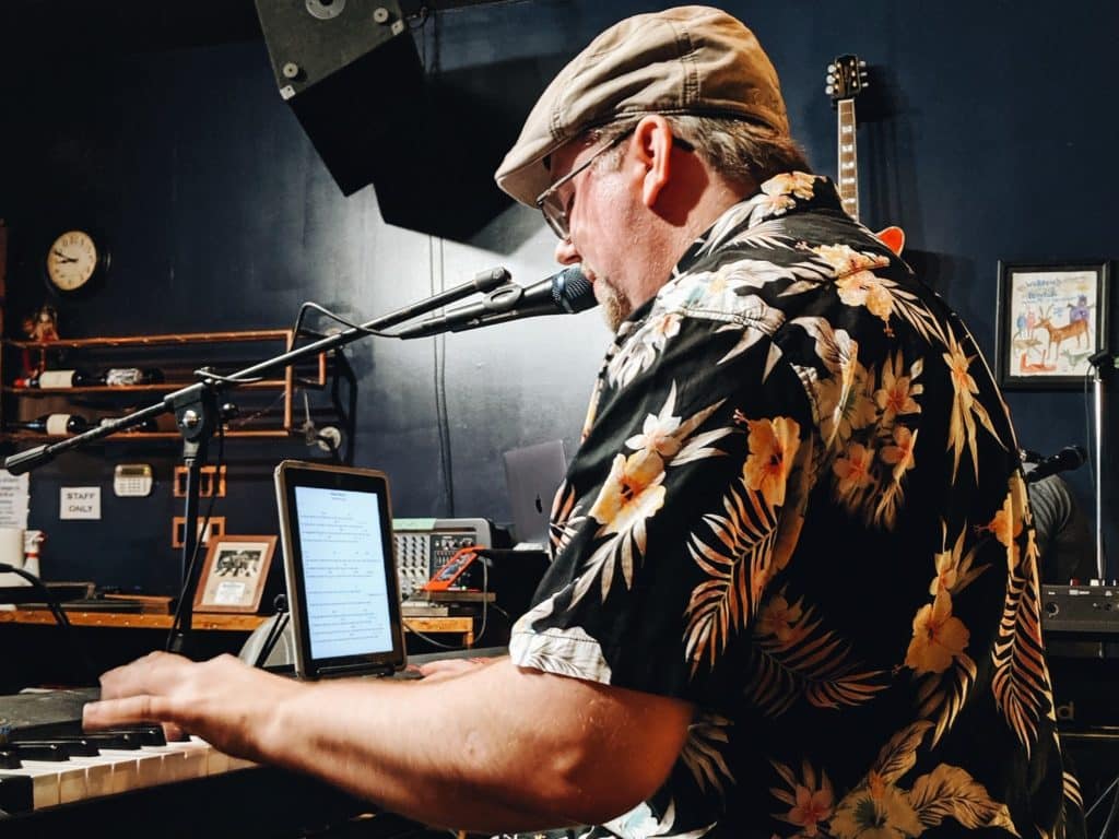 A man with a Hawaiian shirt plays the keyboard in a soundroom with a mic at his mouth.