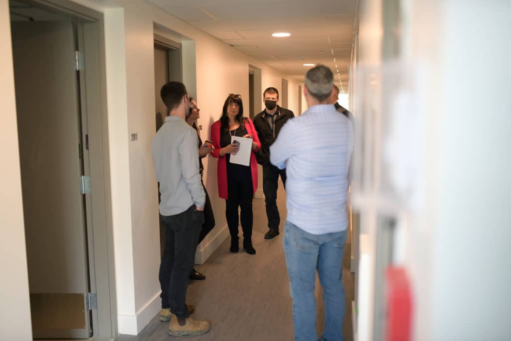 A group of people stand in a hallway of the supportive housing complex on Paddle Road, all looking at Mayor Michelle Staples, who is wearing a bright pink blazer and black pants and seems to be listening to someone speak. 