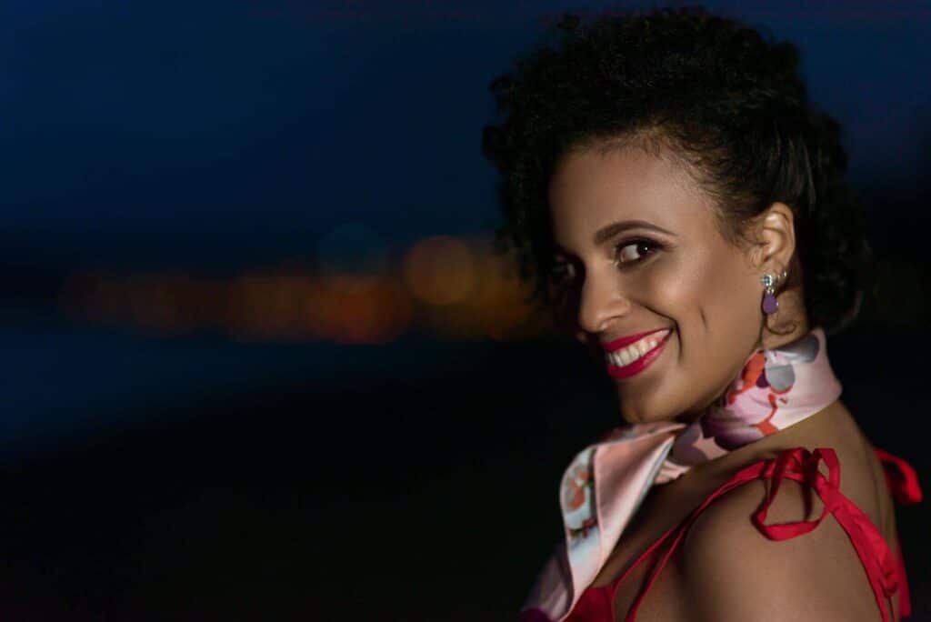 Tania Amaral smiles waring red lipstick and a scarf, with her left profile in view.