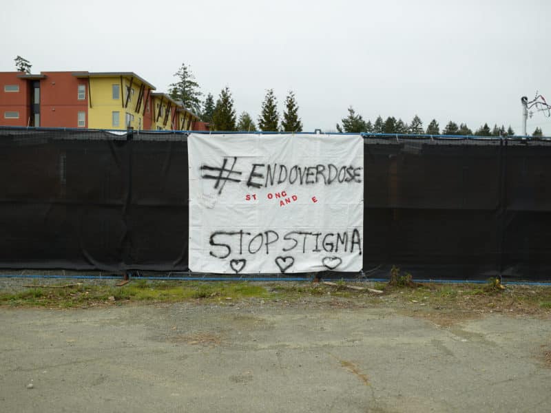 A white plastic sheet hangs on a fence. Spray painted on it is "#End Overdose" and "Stop Stigma" with some spray painted hearts in black and unrecognizeable red sticker letters on the sheet as well.