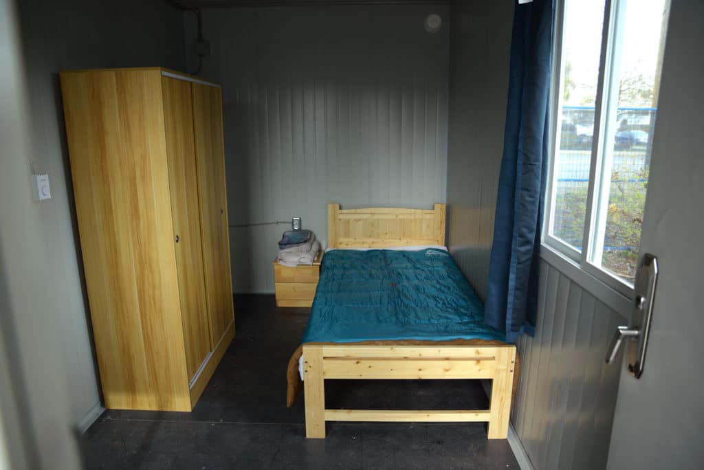 Inside one of the tiny homes at The Village. It is a small, rectangular room. On the right is a single-bed with a light wood bedframe and blue sheets. On the left is a closet that matches the bed frame. In between is a small nightstand. On the far right wall is a window.