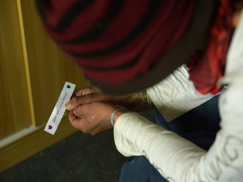 Photo taken from above a man's head. His red and black striped toque is visible in the top half of the photo. The bottom half is filled with his tattooed hands, white long sleeves meeting them, holding a think strip of paper that says "you are loved."