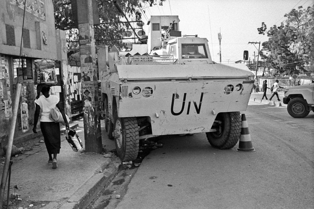 A black and white photo of a Haitian woman walks past a UN truck parked on the sidewalk and half blocking the street.