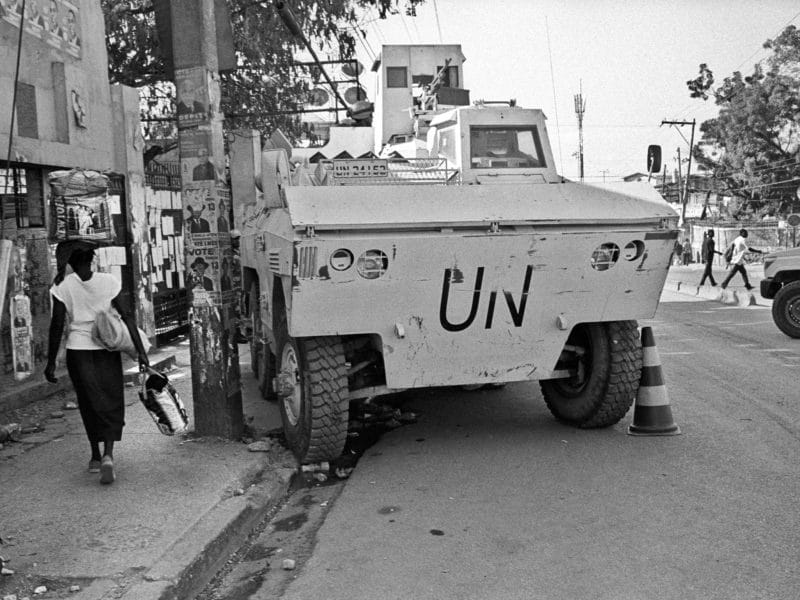 A black and white photo of a Haitian woman walks past a UN truck parked on the sidewalk and half blocking the street.
