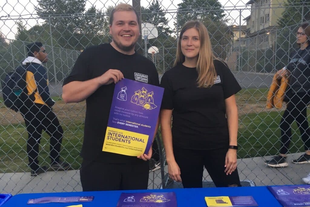 Two people stand in front of a booth and hold up a poster