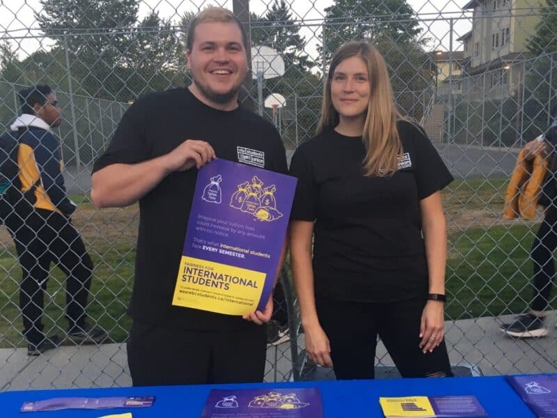 Two people stand in front of a booth and hold up a poster
