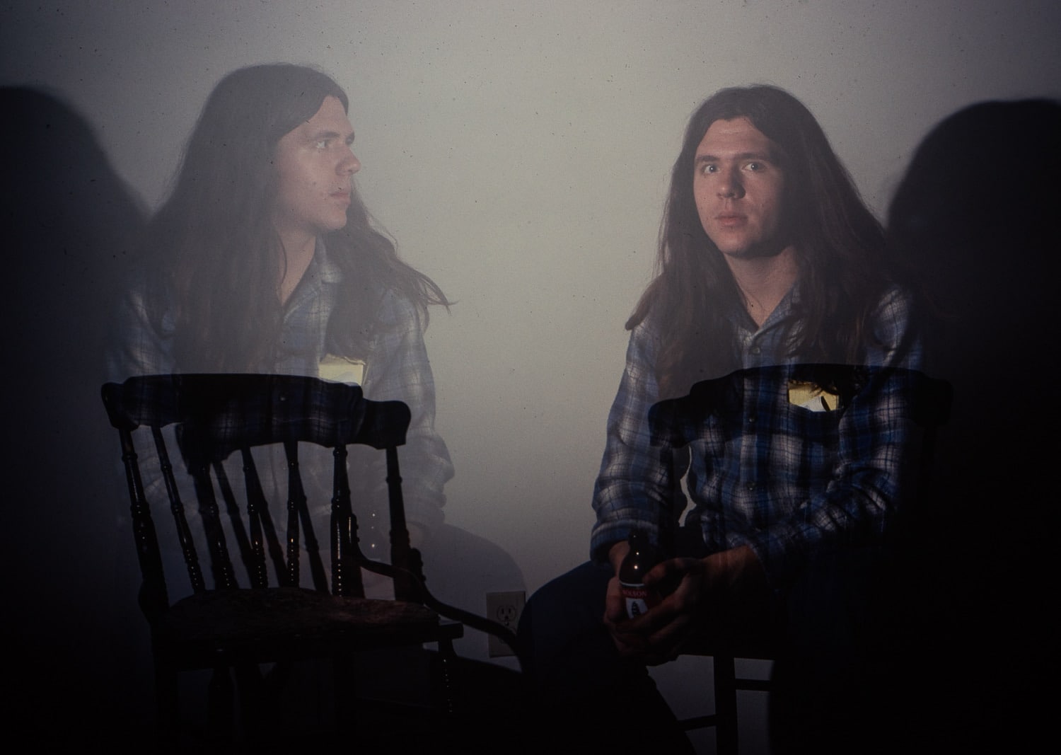 A young Craig Evans with long brown hair sits in a chair facing the camera and holding a beer. The photo has been long exposed so there is a fainter version of Craig sitting in the chair beside the first, looking at first Craig.