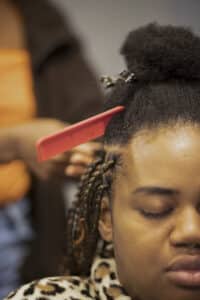 A woman smiles as she combs and braids another Black woman's hair.