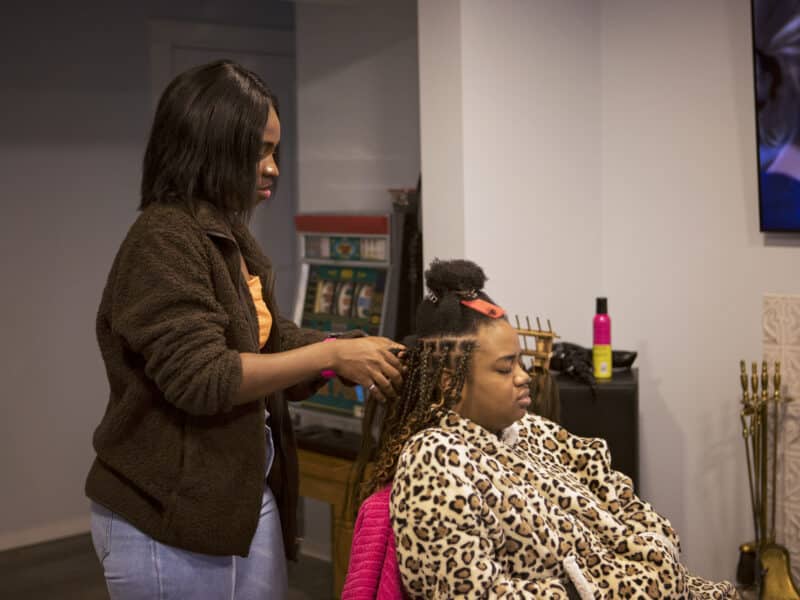 A Black woman standing combs hair away from another Black woman who is sitting.