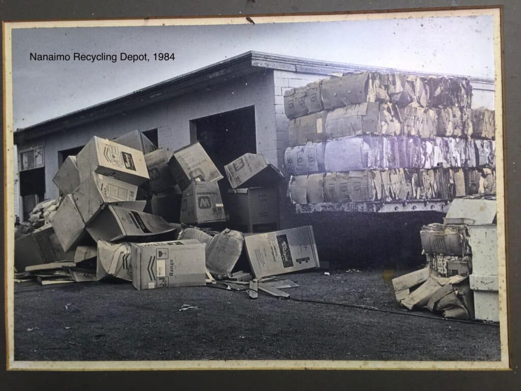 A photo of the original Nanaimo recycling depot, dated 1984. There are multiple bundles of cardboard to the right of the building, and a pile that reaches the top of the door in front.