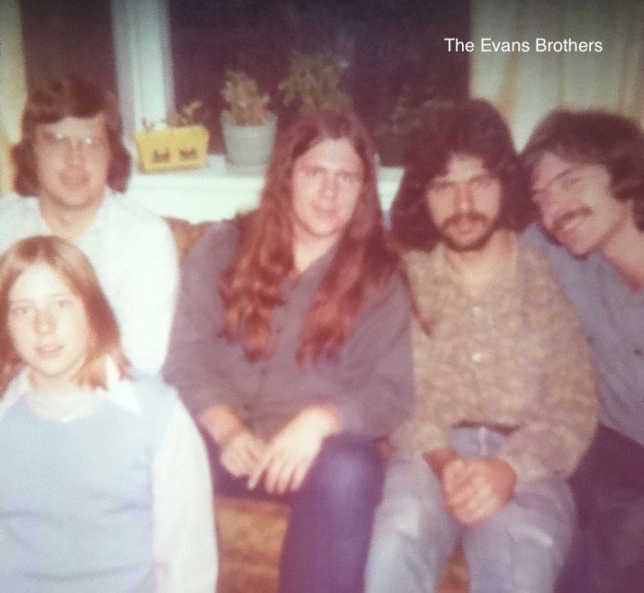 An older polaroid photo of five men at various ages sitting on and in front of a couch. They are all smiling and looking at the camera. Craig Evans sits in the middle with long brown hair.