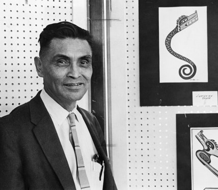 A black and white photo shows a clean shaven man in a suit standing beside frame artwork.
