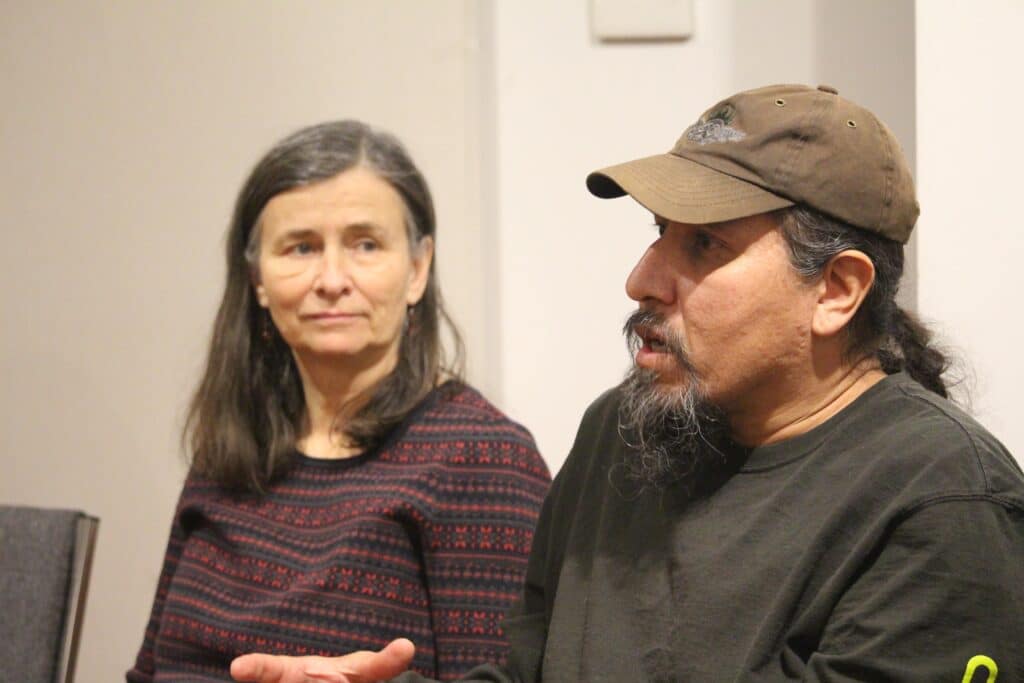 Tim Kulchyski is in the right of the photo, seated and speaking. The photo is taken from the chest up. He wears a sun-faded, black baseball cap and a black long-sleeve. To the left of him in and slightly in the background, out of focus, is Heather Pritchard. She is facing Tim, listening. She wears a black long-sleeve with a red striped pattern on it. Tim is answering questions about solutions for local watersheds.