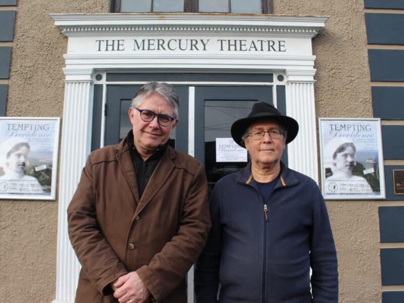 Rien Vesseur and Jim Cleough stand in front of the entrance of the Mercury Theatre. They're side-by-side, standing in front of the doors which say "The Mercury Theatre" above them. Rien wears a brown jacket and glasses, Jim wears a black jacket and black fedora.