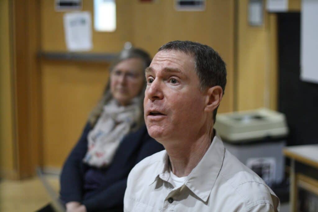 A short-haired man makes an expressive face seated in a white button up shirt with a woman in the background wearing a scarf. 