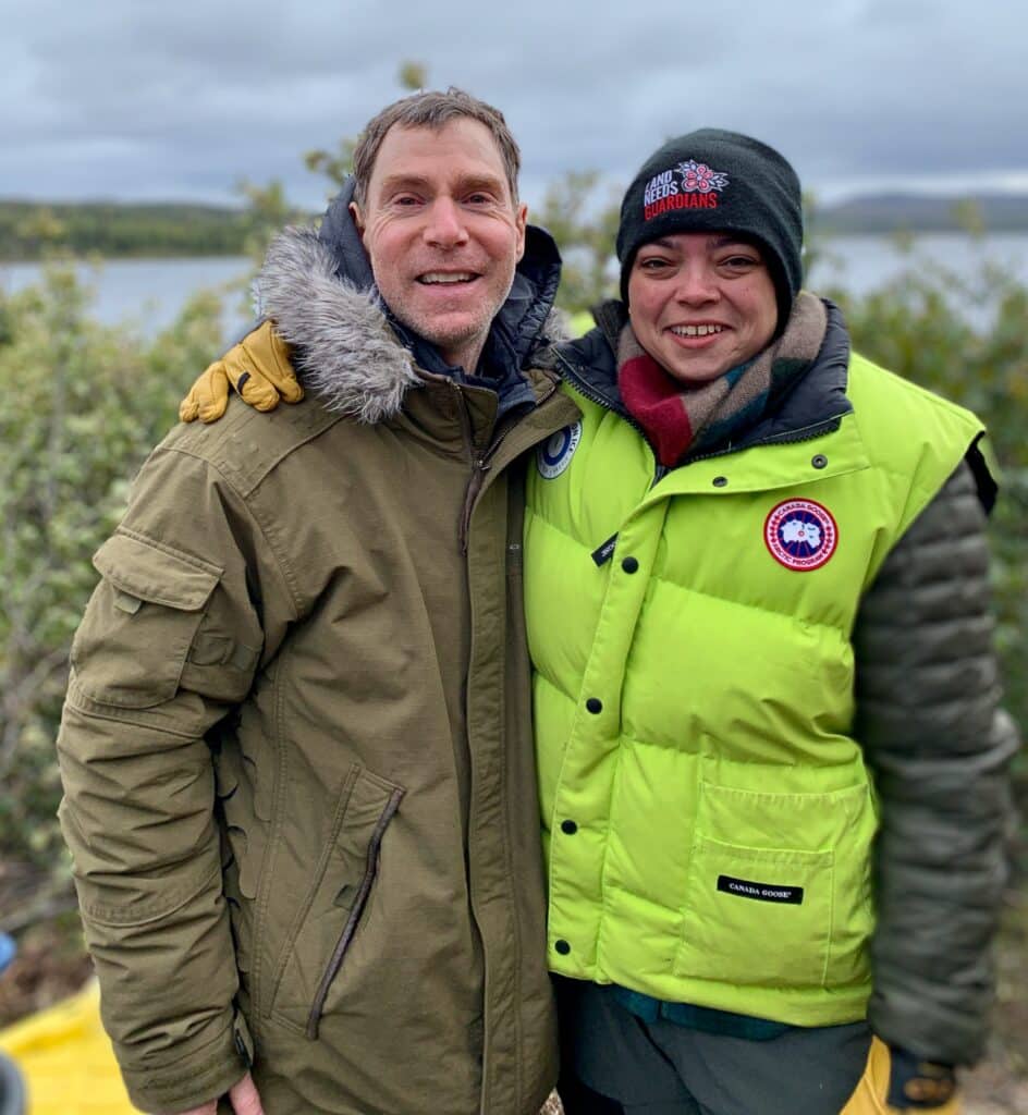 Two people stand side by side in winter coats with a grey stormy background and shrubs.