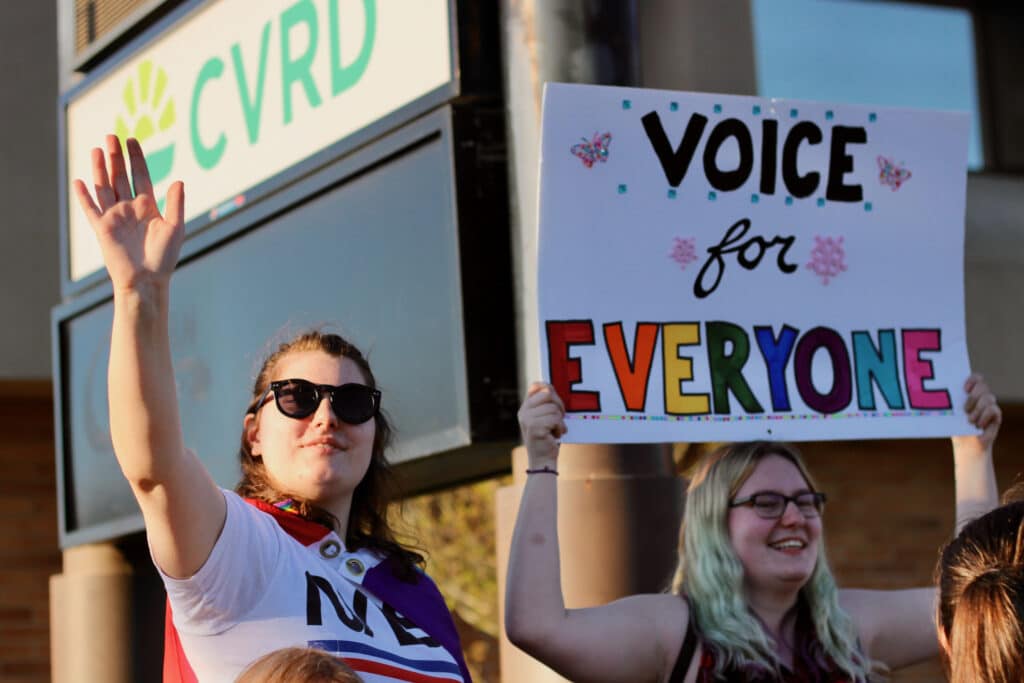 Two youth are pictured in the photo. The one on the left is wearing sunglasses with one hand in the air as if waving and the one on the right holds a large sign above their head that says "Voice for everyone" as they smile.