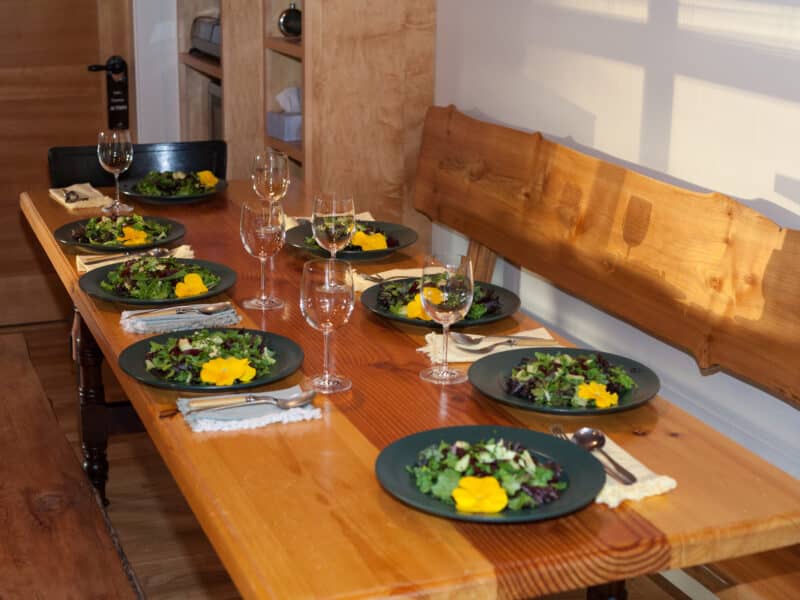 A long wooden table is neatly set with eight colourful plates of salad