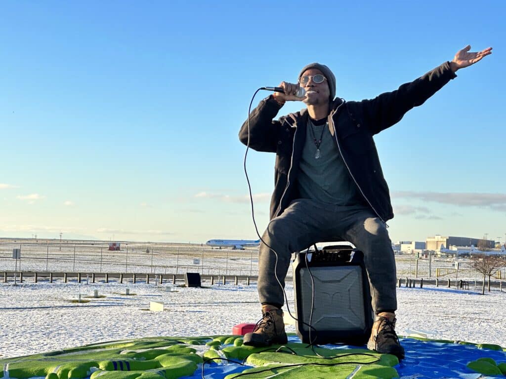 An artist with a mic sits on a speaker at the airport with the runway covered in a light blanket of snow and open blue skies.