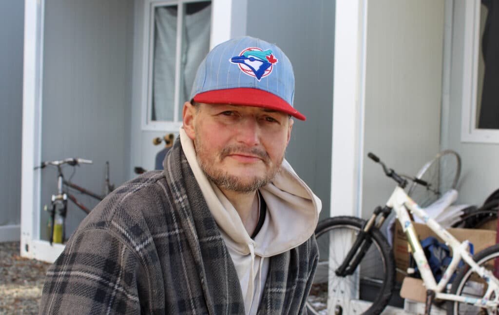 Jeffrey “Disco” Bell is pictured, from shoulders up, sitting in front of a temporary housing site where he lives. He is wearing a Blue Jays baseball cap, hoodie and plaid jacket. There is a white bike in the background as well.