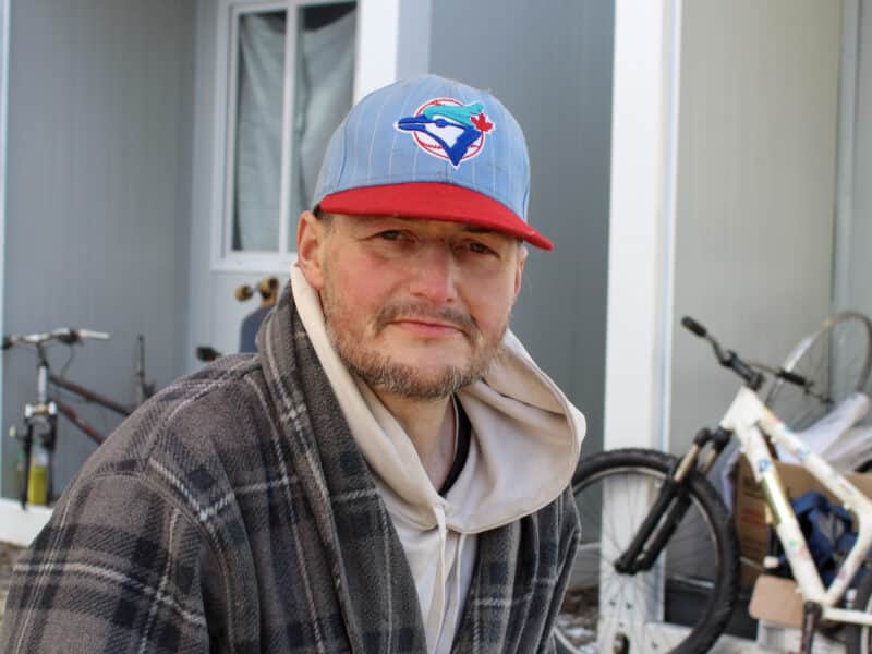 Jeffrey “Disco” Bell is pictured, from shoulders up, sitting in front of a temporary housing site where he lives. He is wearing a Blue Jays baseball cap, hoodie and plaid jacket. There is a white bike in the background as well.