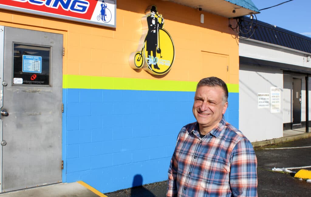 Will Arnold stands, facing the camera and smiling, with his shop Experience Cycling in the background. He wears a plaid shirt.