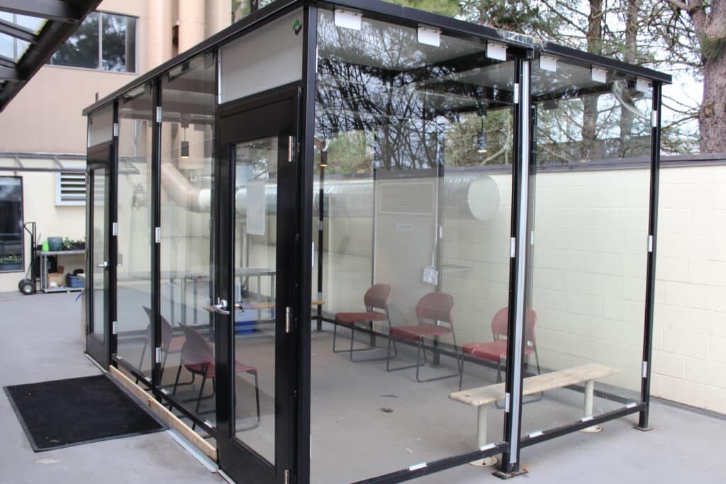 The inhalation area at the overdose prevention site located at the wellness centre is pictured. It is a large rectangular glass box that looks like a bus stop. Three chairs are placed against the walls on the long side of the box and two small benches are against the wall on the short side of the box. An exhaust pipe is seen connected to the back of the box.