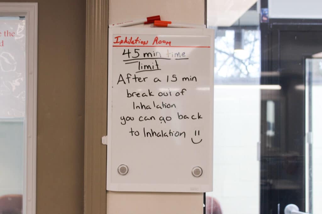 A whiteboard sign at the wellness centre hangs on the wall and reads "Inhalation room 45 min time limit After a 15 min break out of inhalation you can go back to inhalation" with a drawn-on smiley face after the wording.
