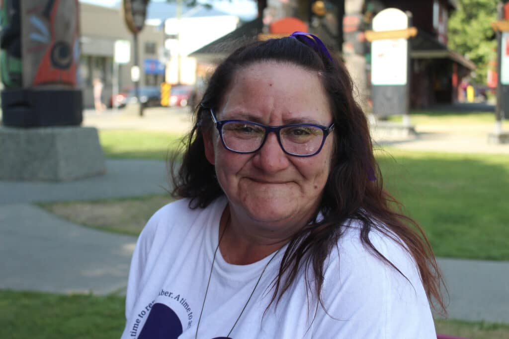 Tracy, a peer worker with the Cowichan Community Action Team and client of the wellness centre and Overdose Prevention Site is pictured, facing the camera and smiling from the shoulders up. She is seated outdoors, with grass and sidewalks of downtown Duncan behind her. She wears glasses, a t-shirt and a ribbon in her hair which is in a partial up-do.