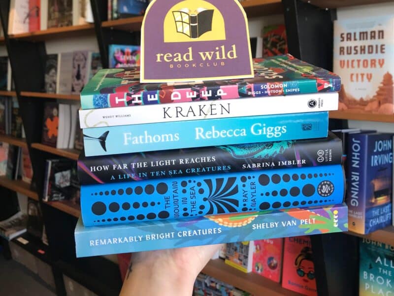 A hand holds up a collection of ocean themed books. In the background are shelves of books. The ocean themed books include The Deep by Rivers Solomon, Kraken by Wendy Williams, Fathoms by Rebecca Giggs, How Far the Light Reaches by Sabrina Imbler, The Mountain in the Sea by Ray Nayler, and Remarkably Bright Creatures by Shelby van Pelt