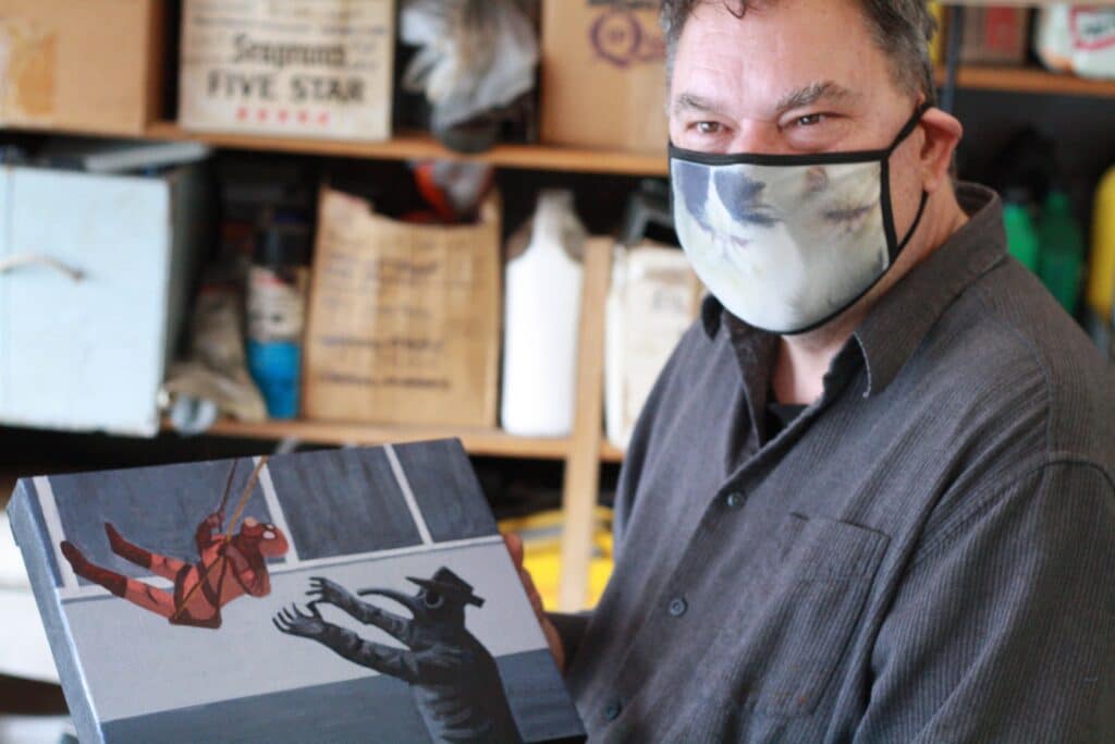 Artist Rod Corraini wearing a mask and holding up one of his plague family paintings as he looks to the left of the camera and seems to be smiling at someone.