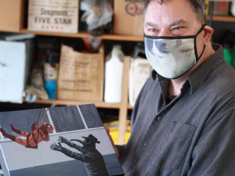 Artist Rod Corraini wearing a mask and holding up one of his plague family paintings as he looks to the left of the camera and seems to be smiling at someone.